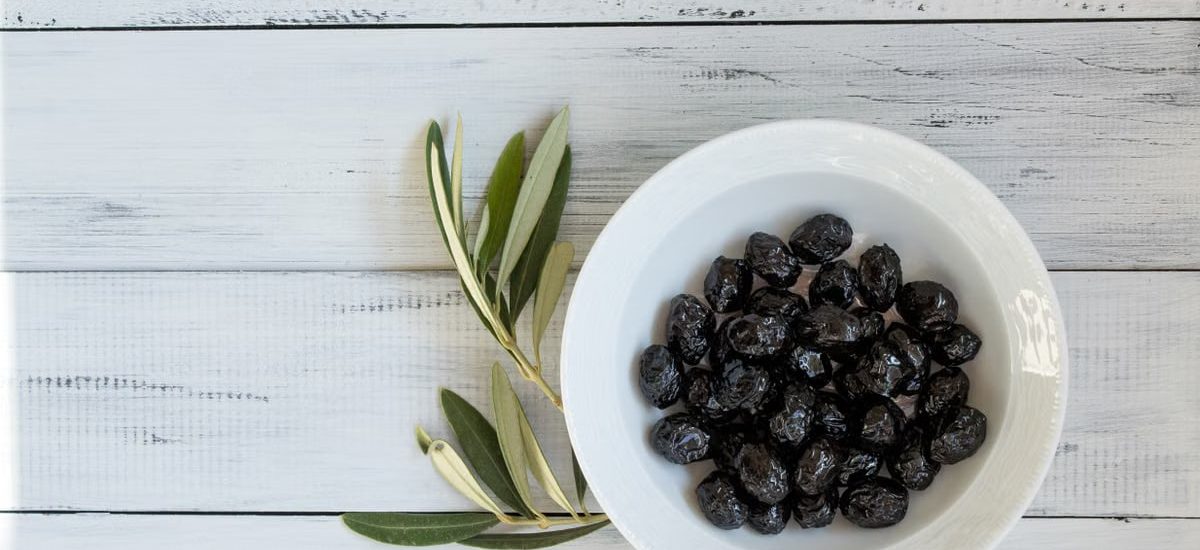 A bowl of black olives on a rustic white wooden surface with olive leaves. Perfect for culinary themes.