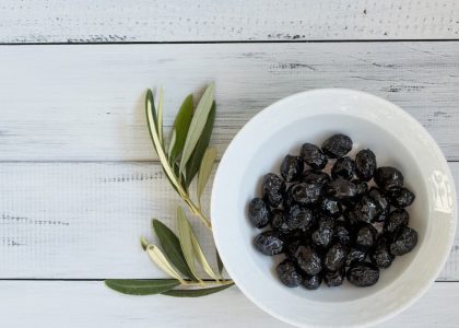 A bowl of black olives on a rustic white wooden surface with olive leaves. Perfect for culinary themes.