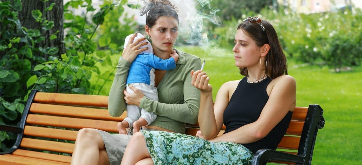 A girl smokes a cigarette on a park bench and disturbs her friend, a young mother and her baby, with her smoke – an appeal for a little more attention and awareness when smoking outdoors. Eine junge Frau raucht auf einer Parkbank eine Zigarette und belästigt mit ihrem Rauch ihre Freundin, eine junge Mutter mit ihrem Baby – ein Aufruf zu etwas mehr Rücksicht und Achtsamkeit beim Rauchen im Freien.